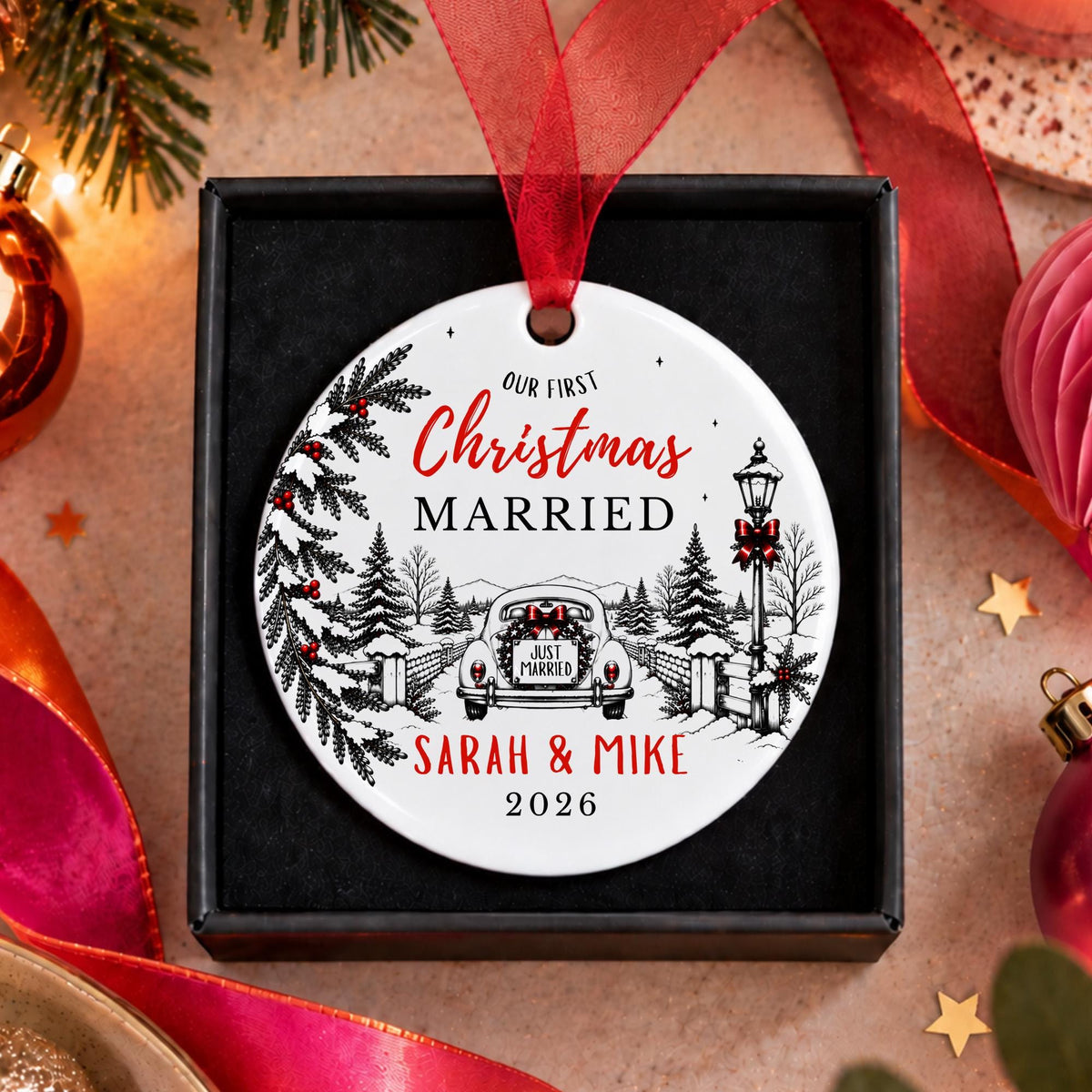 A personalized round Christmas ornament displayed in a black gift box, featuring a winter village illustration and the text 'Our First Christmas Married Sarah & Mike 2026' with a red ribbon hanging above.