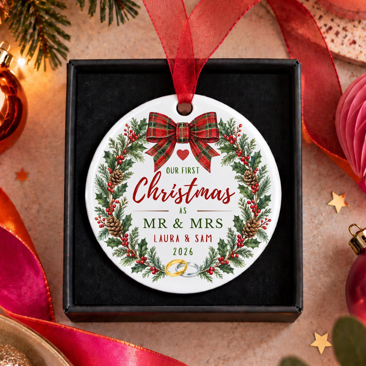 A personalized round Christmas ornament displayed inside a black gift box, featuring a festive wreath design with a red plaid bow and custom text reading 'Our First Christmas as Mr. & Mrs.
