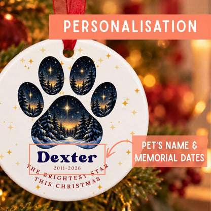 Personalized Christmas pet memorial ornament featuring a paw print with starry night sky forest scenes, the name 'Dexter' and memorial dates '2011-2026', and the phrase 'THE BRIGHTEST STAR THIS CHRISTMAS'. The ornament is shown hanging in a festive blurred holiday setting with text overlays highlighting personalization.