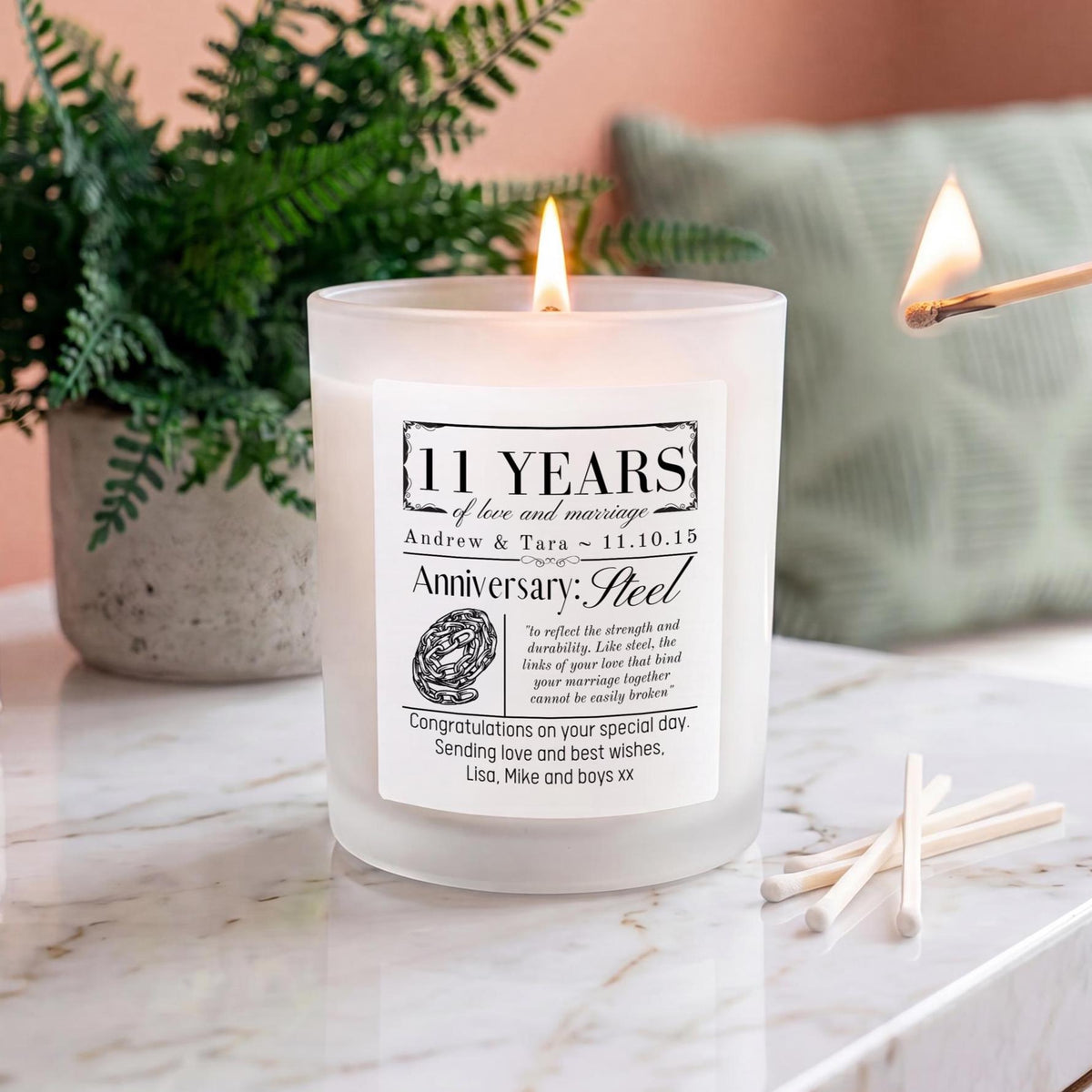 A frosted glass candle with a lit flame sits on a marble surface, styled with a potted fern, matches, and a lit match in the background.