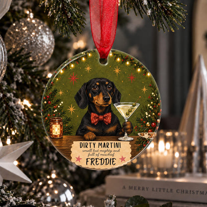 A personalized round Christmas ornament hanging on a decorated Christmas tree. The ornament features a black and tan dachshund holding a martini glass, with the text 'DIRTY MARTINI small time martini man full of mischief FREDDIE' on a green holiday background with warm string lights.