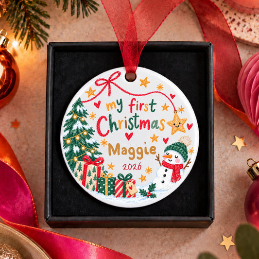A personalized round Christmas ornament displayed in a black gift box, featuring festive illustrations of a Christmas tree, presents, a snowman, and the text 'my first Christmas Maggie 2026' with a red ribbon loop at the top.