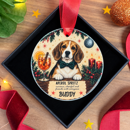 A personalized round Christmas ornament featuring a beagle dog wearing a green bow tie, with festive illustrations of an aperol spritz, candles, holly, and stars. The ornament is displayed inside a black gift box on a wooden surface, decorated with red ribbons and holiday-themed props.