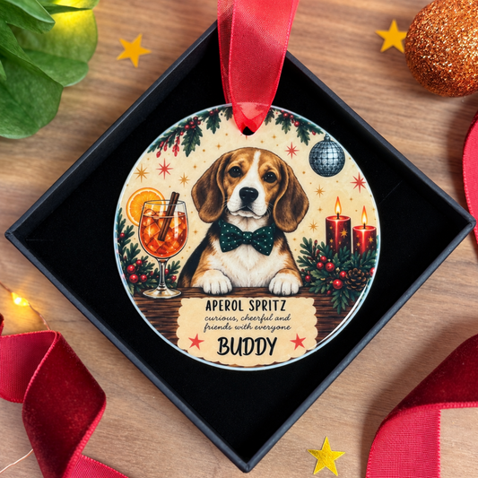 A personalized round Christmas ornament featuring a beagle dog wearing a green bow tie, with festive illustrations of an aperol spritz, candles, holly, and stars. The ornament is displayed inside a black gift box on a wooden surface, decorated with red ribbons and holiday-themed props.