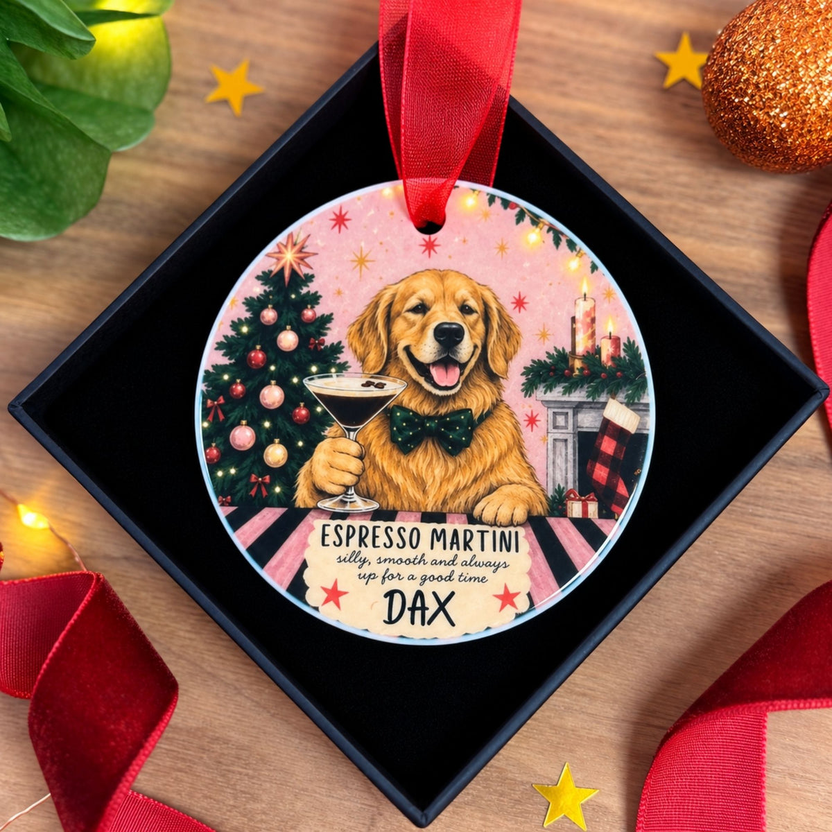 Personalised Golden Retriever Christmas ornament displayed inside a black presentation gift box, featuring a smiling golden dog wearing a green bow tie and holding an Espresso Martini. The ceramic hanging decoration shows a decorated Christmas tree, festive fireplace, stockings and candles, with the personalised name Dax beneath the illustration and a deep red hanging ribbon.