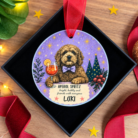 A round personalized Christmas ornament displayed inside a black gift box on a wooden surface, decorated with a brown dog illustration, a purple starry background, a drink, Christmas trees, and the text 'APEROL SPRITZ happy, bubbly and friends with everyone LOKI'.