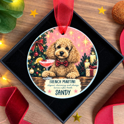 A round personalized Christmas ornament featuring a fluffy golden-brown poodle dog sitting at a table with a cocktail glass, surrounded by a decorated Christmas tree, candles, and gifts. The ornament is displayed inside a black gift box on a wooden surface with festive red ribbons and star decorations.