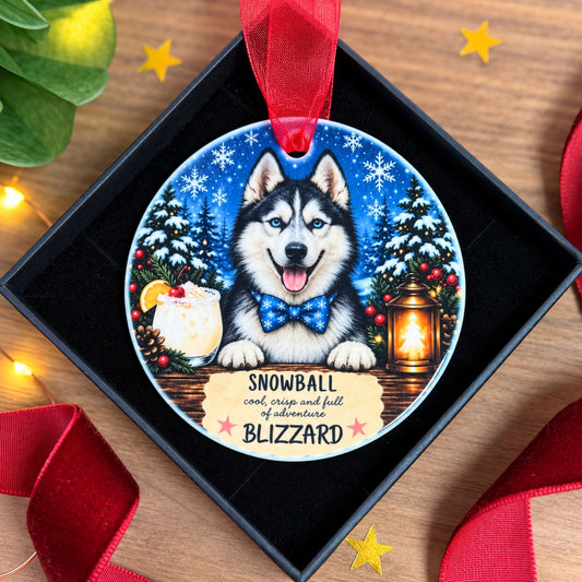 A round Christmas ornament featuring a painted husky dog with the text 'SNOWBALL and crisp and full of adventure BLIZZARD,' displayed inside a black gift box on a wooden surface with red ribbon and holiday decorations.