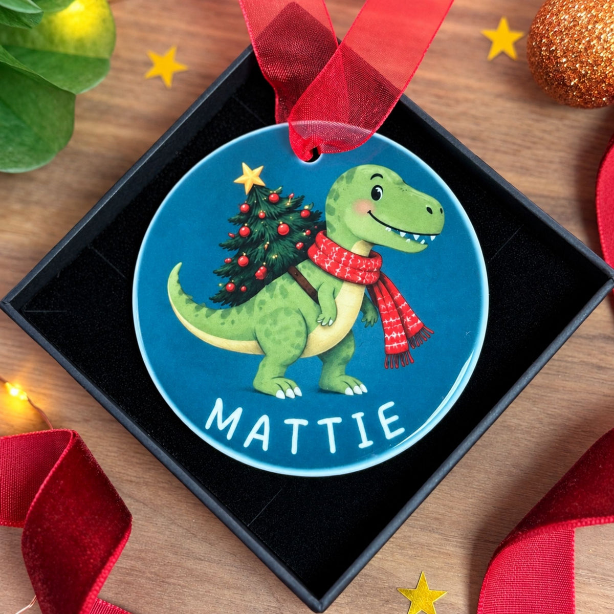 A personalized Christmas ornament shaped like a blue circle with a cartoon green dinosaur carrying a Christmas tree, shown hanging from a red ribbon inside a black gift box on a wooden surface with holiday decorations.