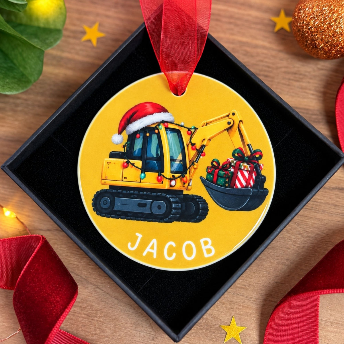 A round personalized Christmas ornament displayed inside a black gift box on a wooden surface. The ornament is yellow and features an illustration of a yellow excavator wearing a Santa hat and holiday lights, with a bucket carrying wrapped presents.