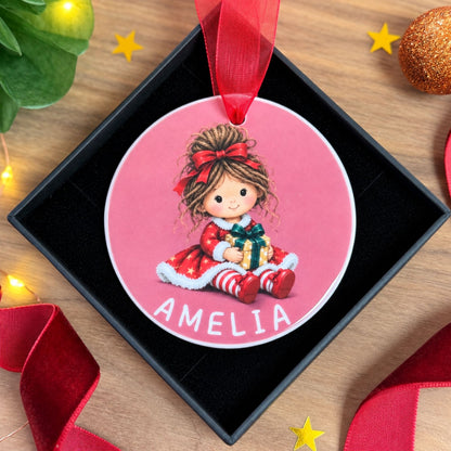 A personalized round Christmas ornament featuring a cartoon girl in a red dress and bow, sitting on a pink background with the name 'AMELIA' at the bottom. The ornament hangs from a red ribbon and is displayed inside a black gift box on a wooden surface with festive decorations.