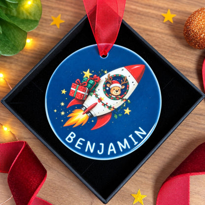 A personalized round Christmas ornament displayed in a black gift box. The ornament is blue with a red ribbon loop, featuring a cartoon reindeer in a white and red rocket ship carrying presents, with the name 'BENJAMIN' printed in white at the bottom.