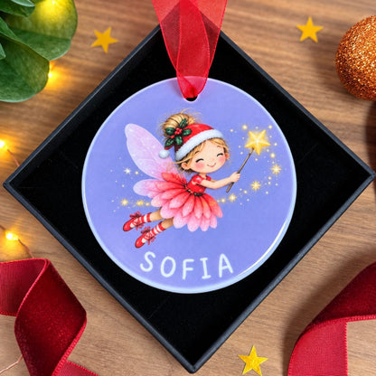 A personalized round Christmas ornament featuring an illustration of a fairy named Sofia on a lavender background, presented in a black gift box with a red ribbon on a decorated wooden surface.