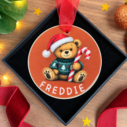 A round Christmas ornament featuring a teddy bear in a red Santa hat and green sweater holding a candy cane, with the name 'FREDDIE' printed at the bottom. The ornament hangs from a red ribbon inside a black gift box on a wooden surface with holiday decorations around it.