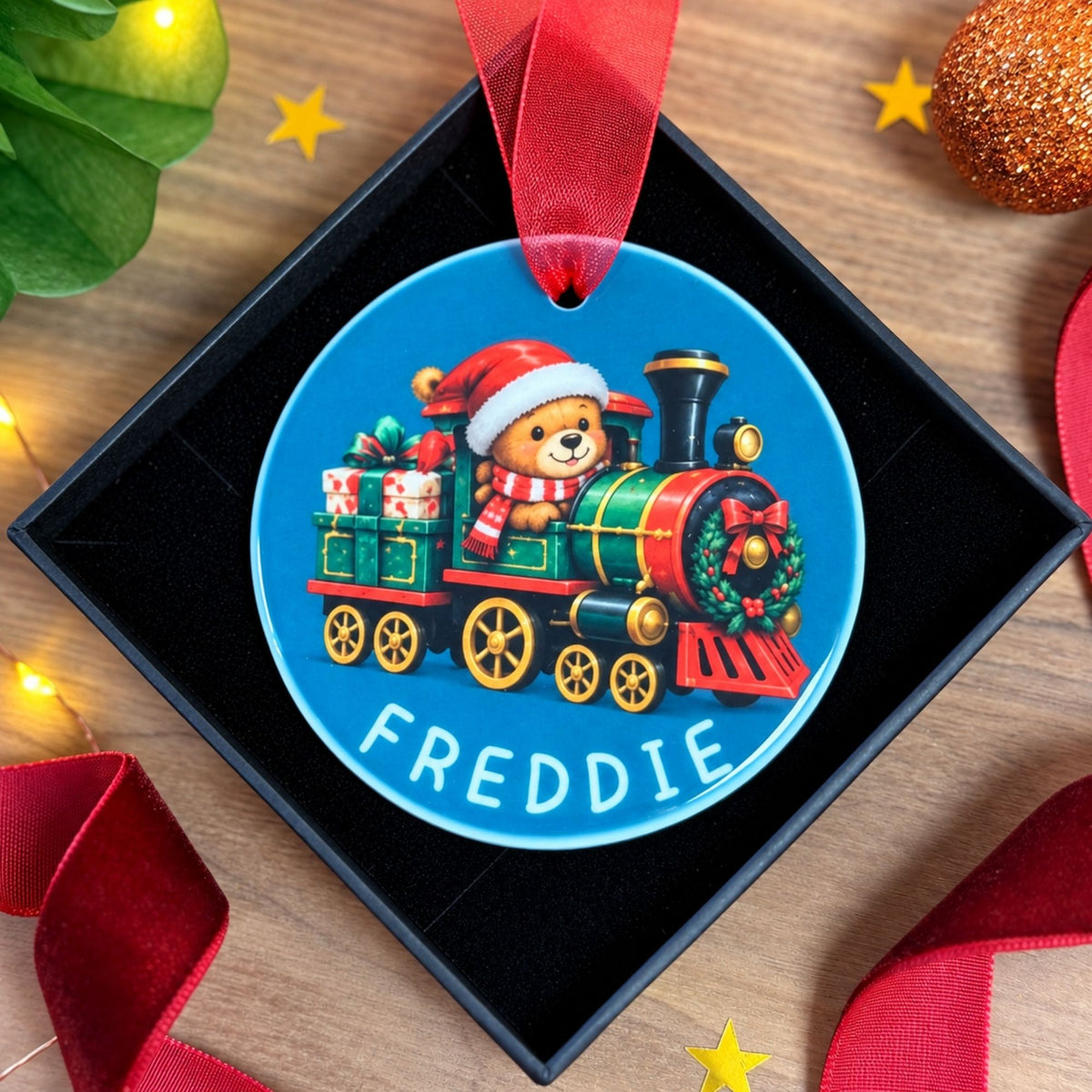 A personalized Christmas ornament shaped like a blue circular train car, displayed inside a black gift box. The ornament features a teddy bear driving a festive green and red train with the name 'FREDDIE' printed in white at the bottom.
