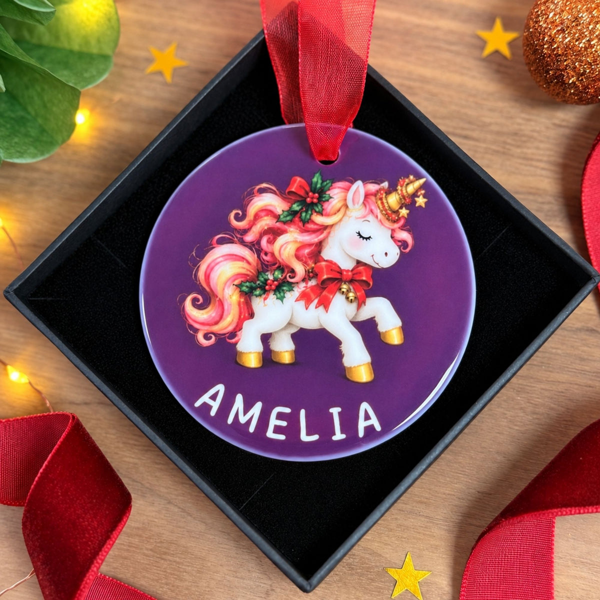 A personalized round Christmas ornament featuring a white unicorn with a pink and orange mane, gold hooves, and red bows, set against a purple background with the name 'AMELIA' in white. The ornament hangs from a red ribbon inside a black gift box on a wooden surface decorated with red ribbons and small yellow stars.