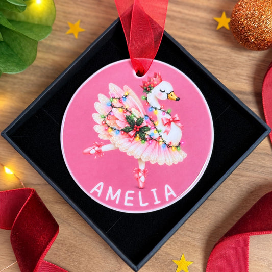 A personalized round Christmas ornament featuring a white swan ballerina in a pink tutu with colorful string lights, hanging in a black gift box on a wooden surface with festive decorations.