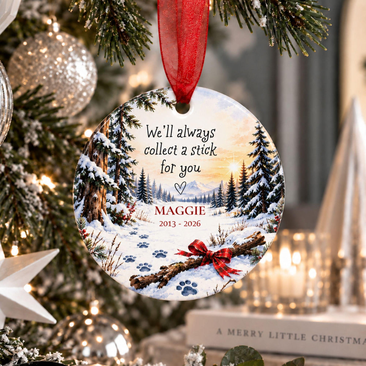 A personalized round Christmas ornament hanging on a decorated tree. The ornament features a snowy forest scene with pine trees, blue paw prints, a red ribbon bow, and the text 'We'll always collect a stick for you' above the name 'MAGGIE 2013 - 2026'.