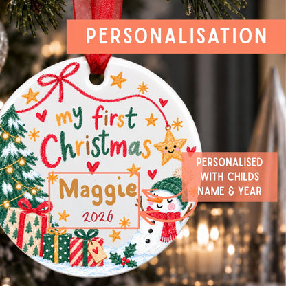 A round white Christmas ornament with a red hanging ribbon, decorated with festive illustrations and the text 'my first Christmas Maggie 2026'. The ornament is shown hanging in front of a blurred Christmas tree and lights, with orange text banners reading 'PERSONALISATION' and 'PERSONALISED WITH CHILD'S NAME & YEAR'.