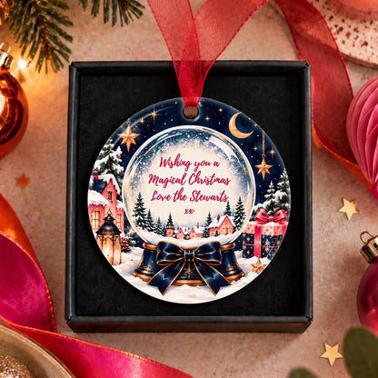 A round Christmas ornament displayed inside a black gift box, featuring a festive winter village scene with bells, stars, and a snow globe motif. The ornament has a red hanging ribbon and is surrounded by holiday decorations like ribbons, ornaments, and pine branches on a warm beige surface.