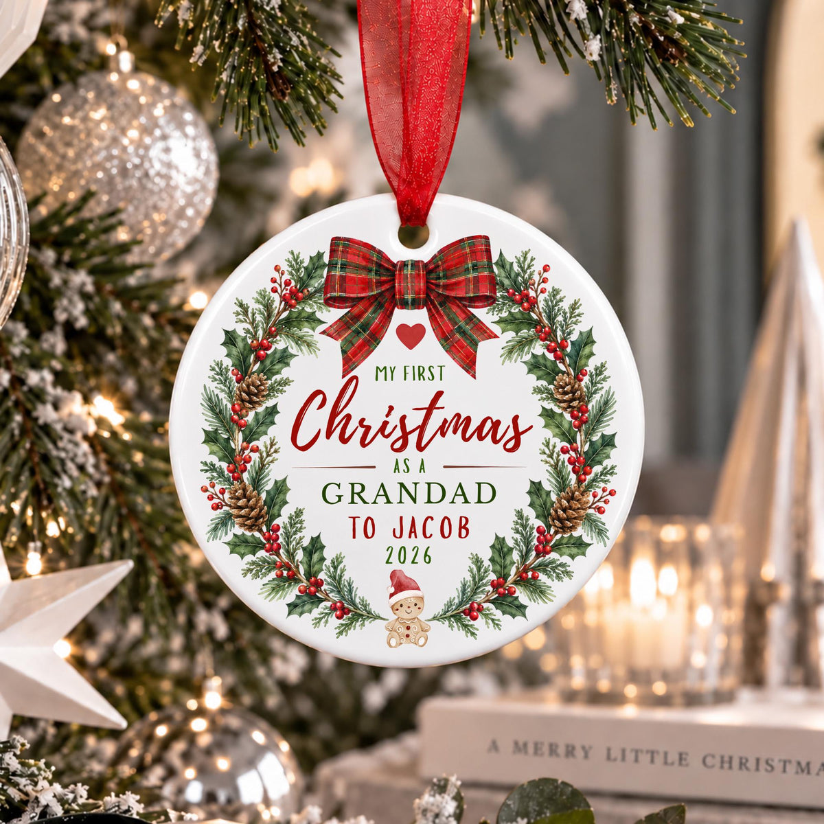 A personalized round Christmas ornament hanging from a red ribbon in front of a decorated Christmas tree. The white ornament features a green holly wreath with red berries, a red plaid bow, and custom text reading 'My First Christmas as a GRANDAD TO JACOB 2026' along with a small baby carriage illustration.