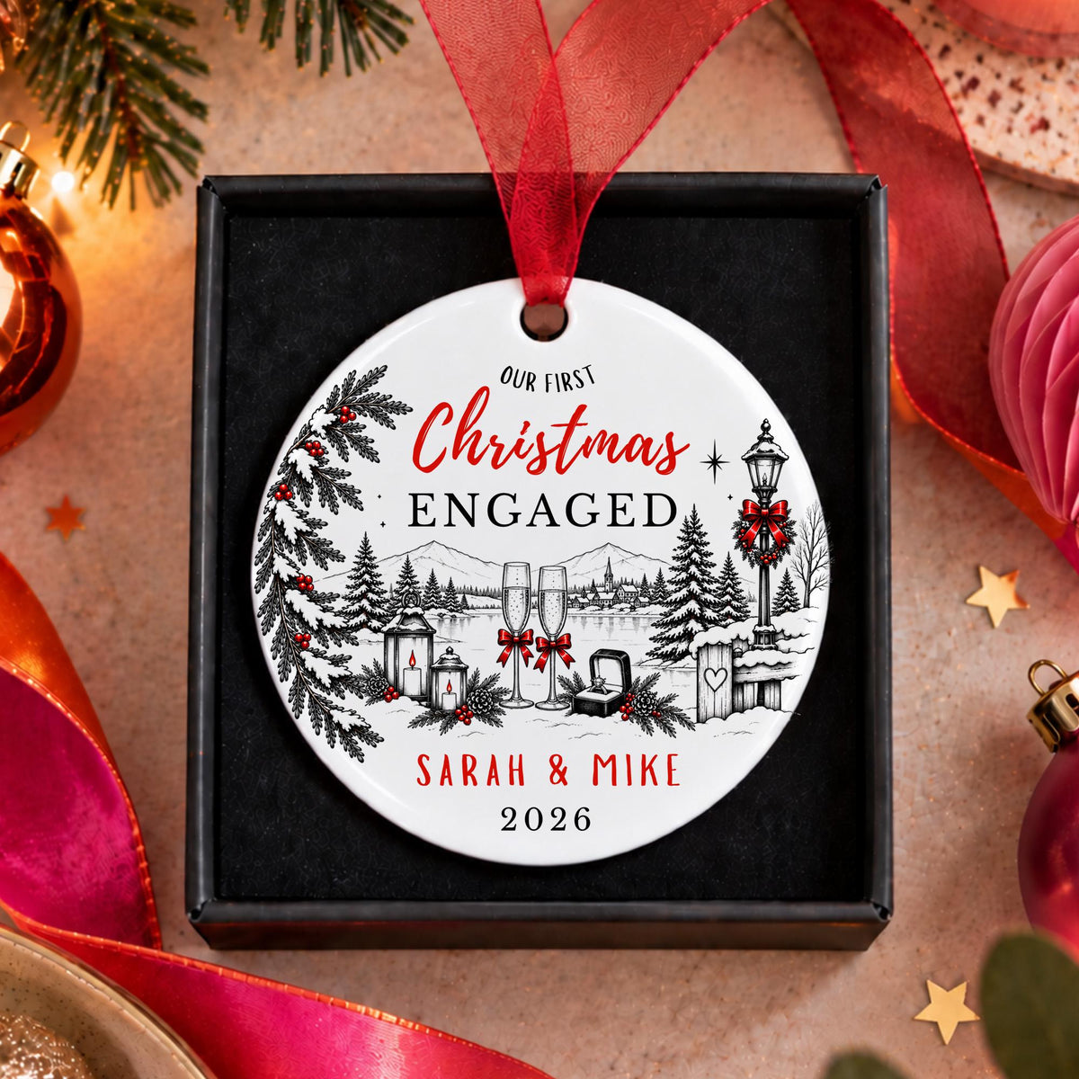 A personalized round Christmas ornament with a red ribbon, displayed in a black gift box. The ornament features a winter village illustration and the text 'OUR FIRST Christmas ENGAGED SARAH & MIKE 2026'.