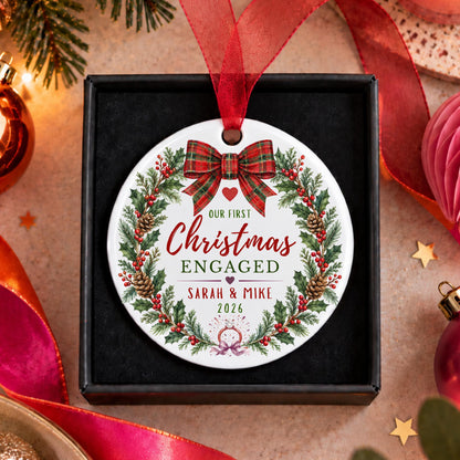 A personalized round Christmas ornament displayed in a black gift box, surrounded by festive holiday decorations including pine branches, red ribbons, and ornaments. The ornament features a white background with a red plaid bow, green pine wreath, red berries, and custom text reading 'Our First Christmas Engaged, Sarah & Mike 2026'