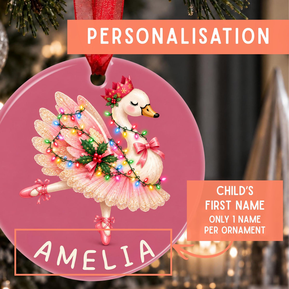 Personalized round pink ballet swan ornament with a red hanging ribbon, decorated with glittery pink tutu, multicolored Christmas lights, holly, and a small red crown. The ornament includes text overlays showing customization options: 'PERSONALISATION' at the top and 'AMELIA' at the bottom, with a note specifying the child's first name is only one per ornament.