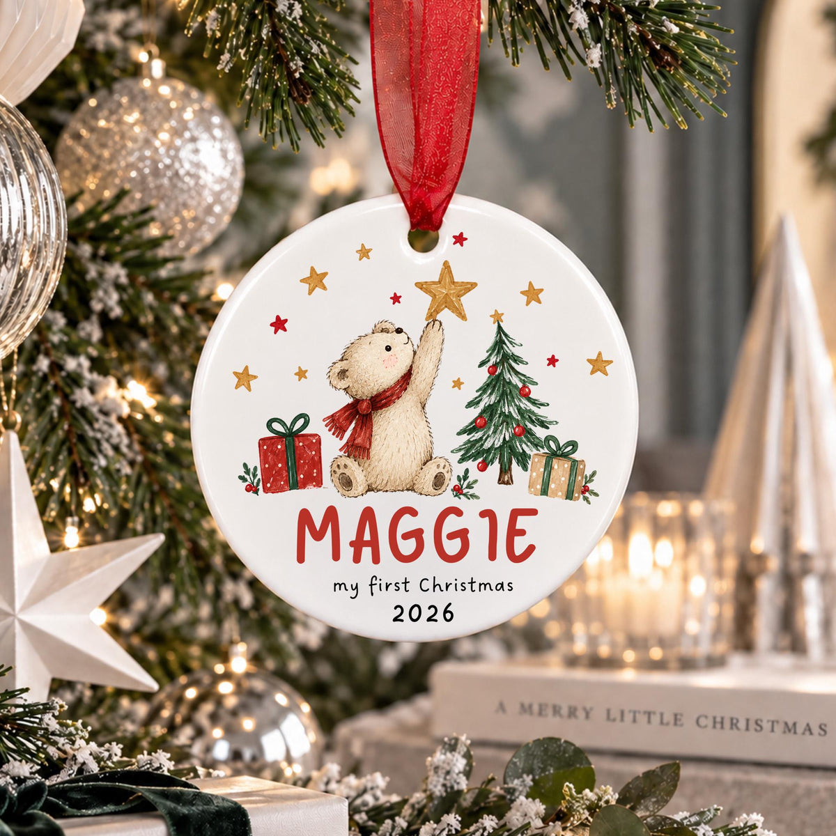 A personalized round Christmas ornament hanging from a red ribbon, featuring an illustration of a teddy bear, a Christmas tree, gifts, and the text 'MAGGIE my first Christmas 2026'. The ornament is displayed in front of a decorated Christmas tree with lights and ornaments.