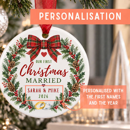 Personalized round Christmas ornament hanging on a tree, featuring a red plaid bow, holly wreath, pinecones, and the text 'Our First Christmas Married Sarah & Mike 2026' with two gold rings. Coral text banners highlight personalization.