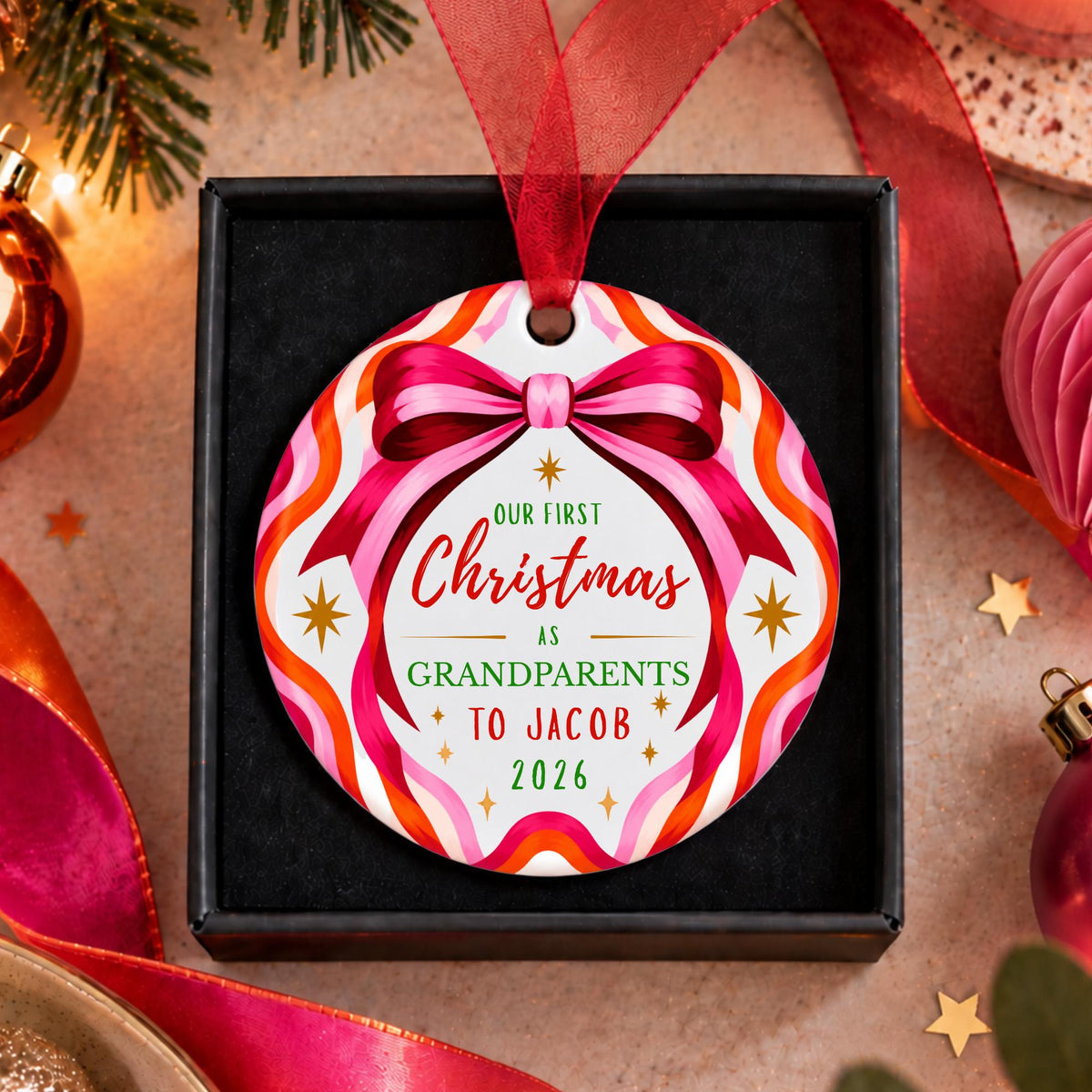 A personalized round Christmas ornament hanging from a red ribbon, displayed in a black gift box. The ornament features a colorful wavy pattern in pink, orange, and white, with a large pink bow graphic and the text 'OUR FIRST Christmas ENGAGED Sarah & Mike 2026'.