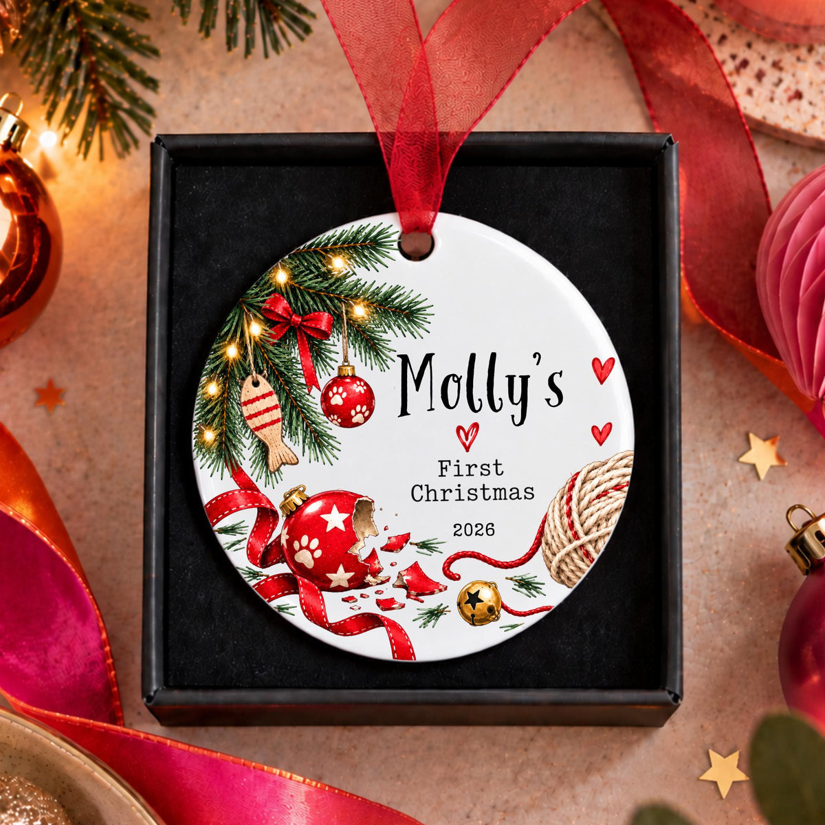 A personalized round Christmas ornament displayed in a black gift box, featuring festive illustrations and the text 'Molly's First Christmas 2026'. The ornament is decorated with red ribbons, pine branches, ornaments, and a star topper, and is surrounded by holiday decorations like pine needles, paper fans, and lights.