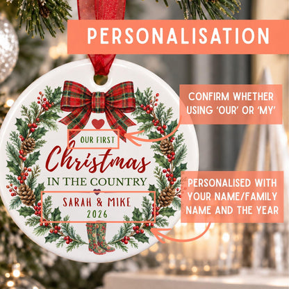 Personalized round Christmas ornament with a red bow and green wreath design, featuring sample text 'Our First Christmas in the Country' and names 'Sarah & Mike 2026', with promotional callouts on a blurred holiday tree background.