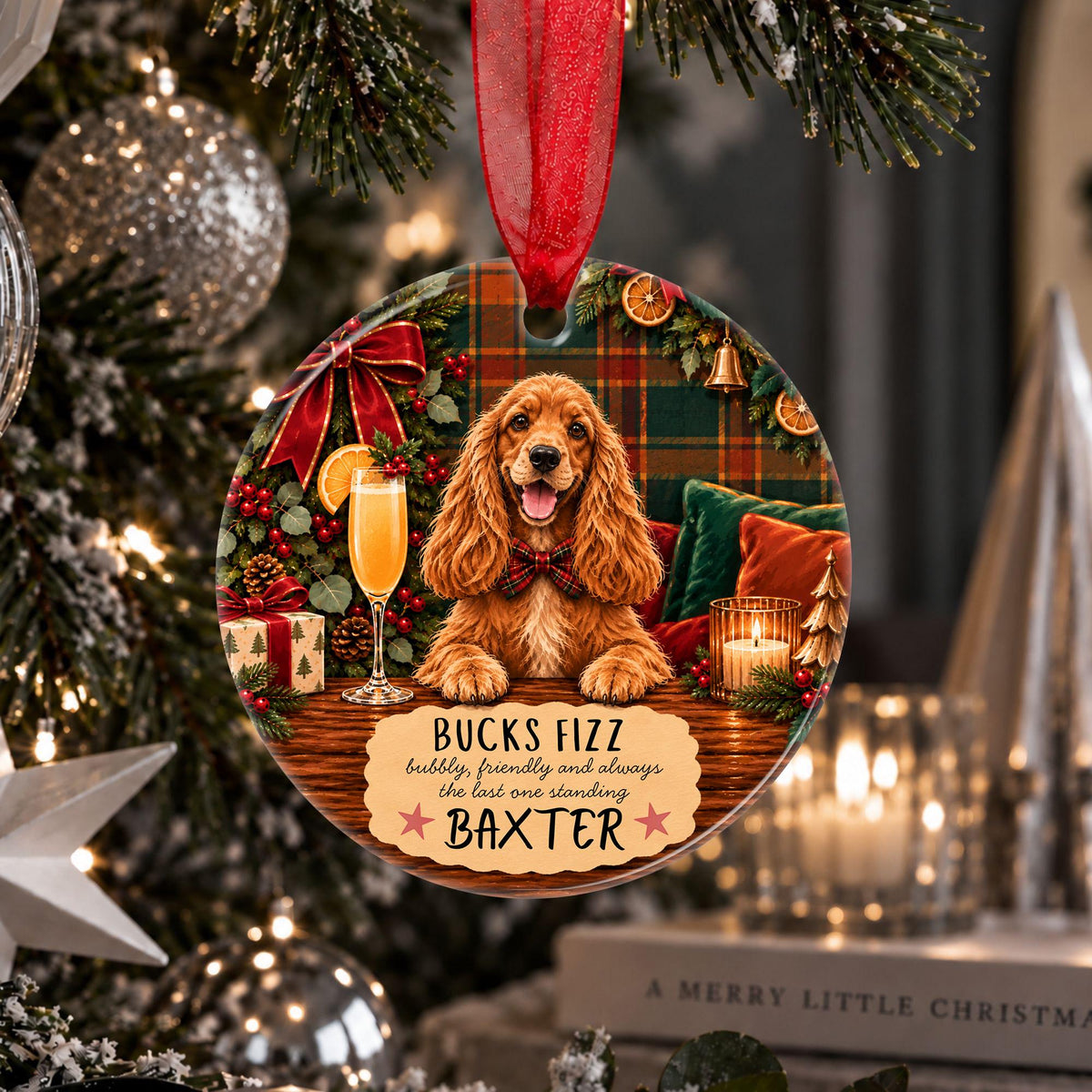 A personalized Christmas ornament shaped like a round wooden plaque hangs from a red ribbon on a decorated Christmas tree. The ornament features a golden-colored dog sitting in front of a holiday scene with a plaid background, a glass of champagne, and the text 'BUCKS FIZZ happily, faithfully and always the last one standing' above the name 'BAXTER'.