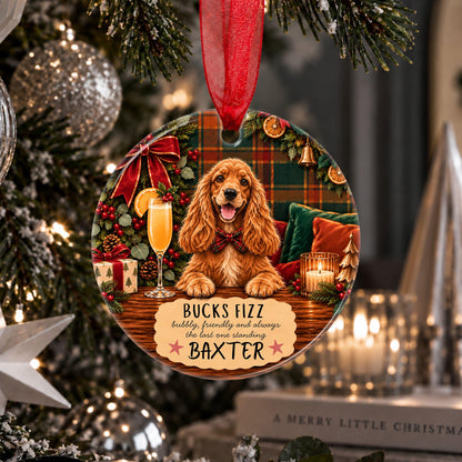 A personalized Christmas ornament shaped like a round wooden plaque hangs from a red ribbon on a decorated Christmas tree. The ornament features a golden-colored dog sitting in front of a holiday scene with a plaid background, a glass of champagne, and the text 'BUCKS FIZZ happily, faithfully and always the last one standing' above the name 'BAXTER'.
