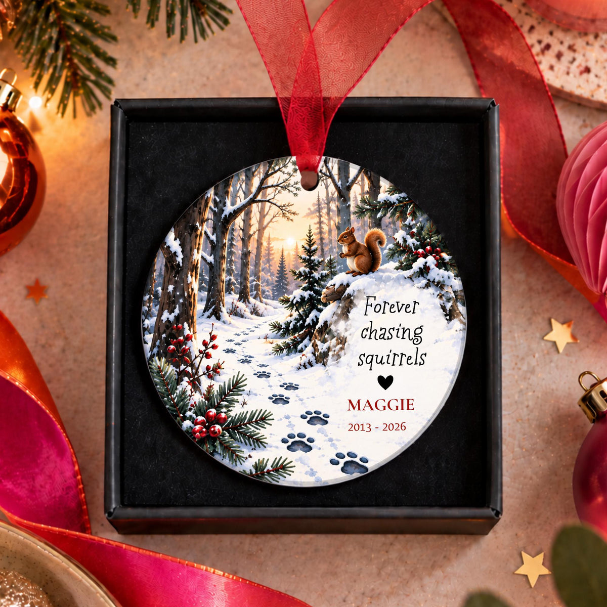 A personalized round Christmas ornament displayed in a black gift box, featuring a winter forest scene with a squirrel, snow, pine branches, and paw prints. The ornament is printed with the text 'Forever chasing squirrels', the name 'MAGGIE', and the years '2013 - 2026'.