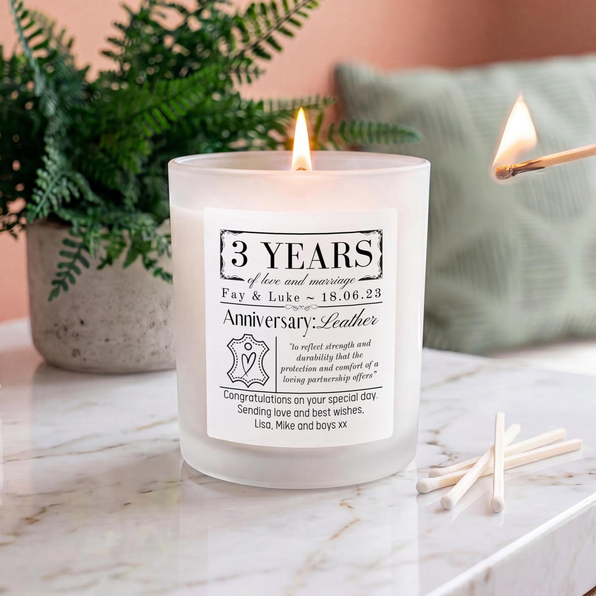 A frosted glass candle with a white label celebrating 15 years anniversary, lit with a visible flame on a marble surface, with a match, another lit flame, and a potted fern in the background.