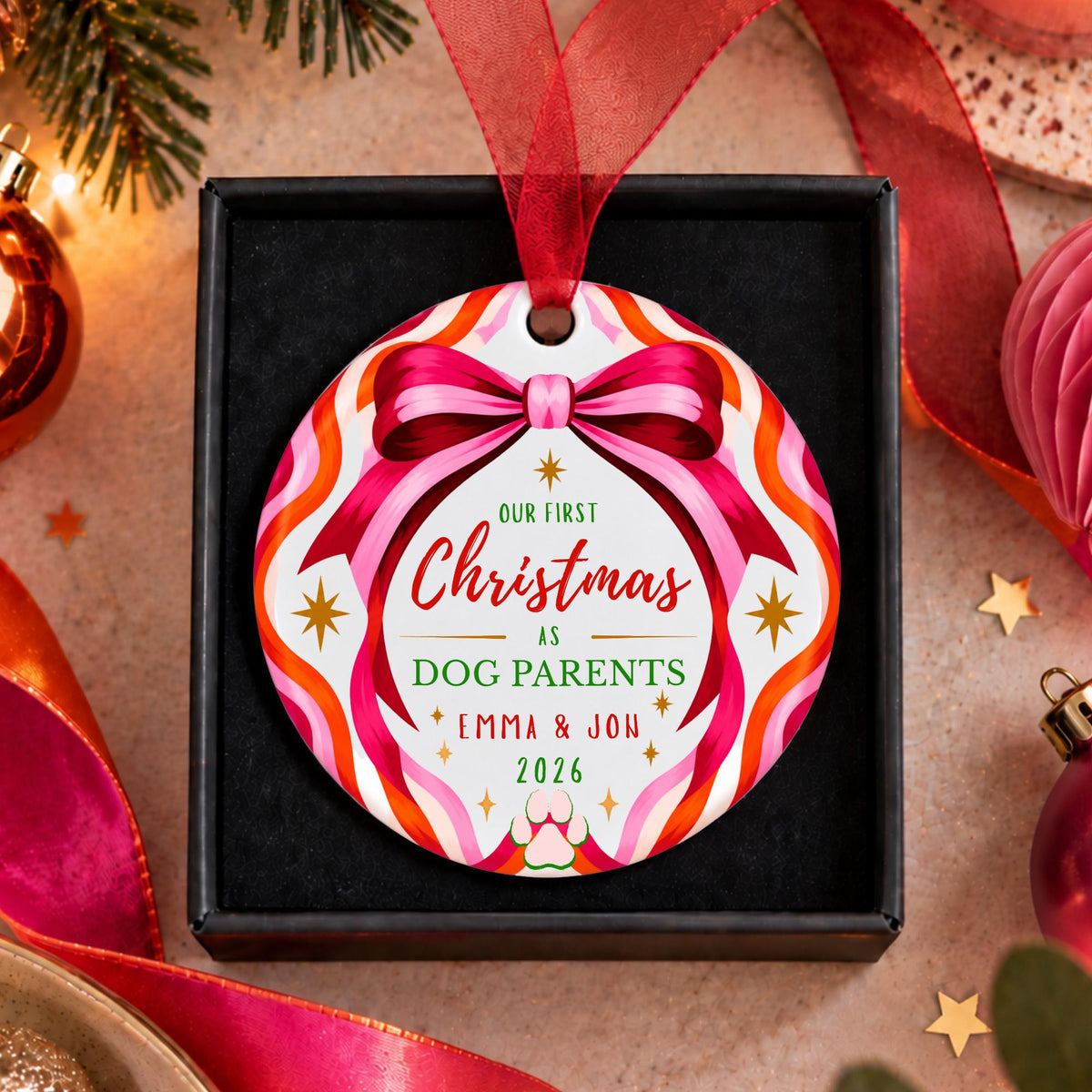 A personalized round Christmas ornament hanging from a red ribbon in a black gift box. The ornament has a colorful retro pattern with pink, orange, and white wavy stripes, a pink ribbon bow graphic, and custom text reading 'Our First Christmas as Dog Parents Emma & Jon 2026'.