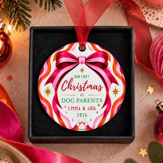 A personalized round Christmas ornament hanging from a red ribbon in a black gift box. The ornament has a colorful retro pattern with pink, orange, and white wavy stripes, a pink ribbon bow graphic, and custom text reading 'Our First Christmas as Dog Parents Emma & Jon 2026'.
