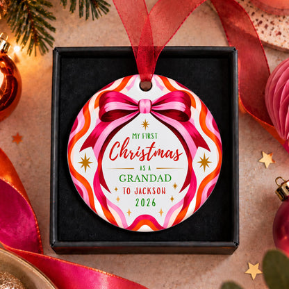 A personalized round Christmas ornament displayed in a black gift box, featuring a colorful pattern of pink, orange, and white wavy stripes with a pink ribbon bow. The center reads 'MY FIRST Christmas AS A GRANDAD TO JACKSON 2026' in red, green, and black text.