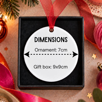 A round white ornament displayed in a black gift box with a red ribbon, printed with dimension labels reading 'ORNAMENT: 7cm' and 'Gift box: 9x9cm'. The scene is styled with Christmas decorations including pine branches, red ribbons, ornaments, and paper flowers on a beige surface.