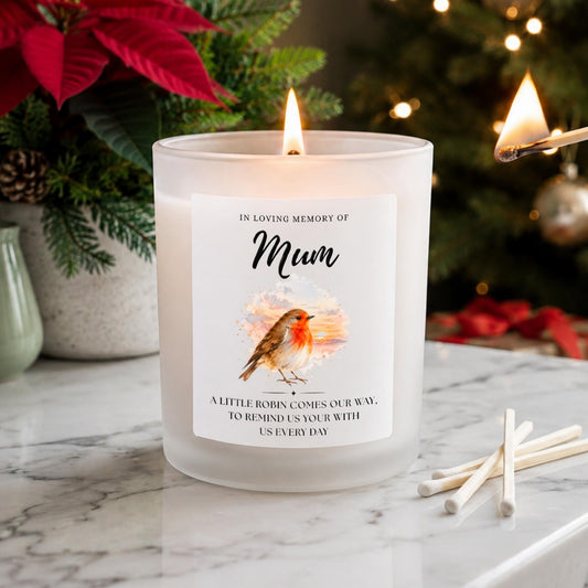 Personalised robin memorial candle in a frosted white glass jar, featuring an In Loving Memory Of Mum label, watercolour robin and remembrance verse. Lit beside Christmas greenery, red poinsettias and white matches.
