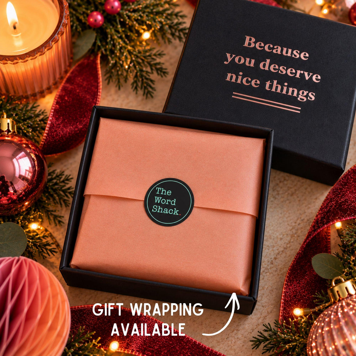 A black gift box with a black lid printed with the phrase 'Because you deserve nice things' sits beside an open box wrapped in peach-colored gift paper. A round black sticker on the wrapped box reads 'The Word Shack.