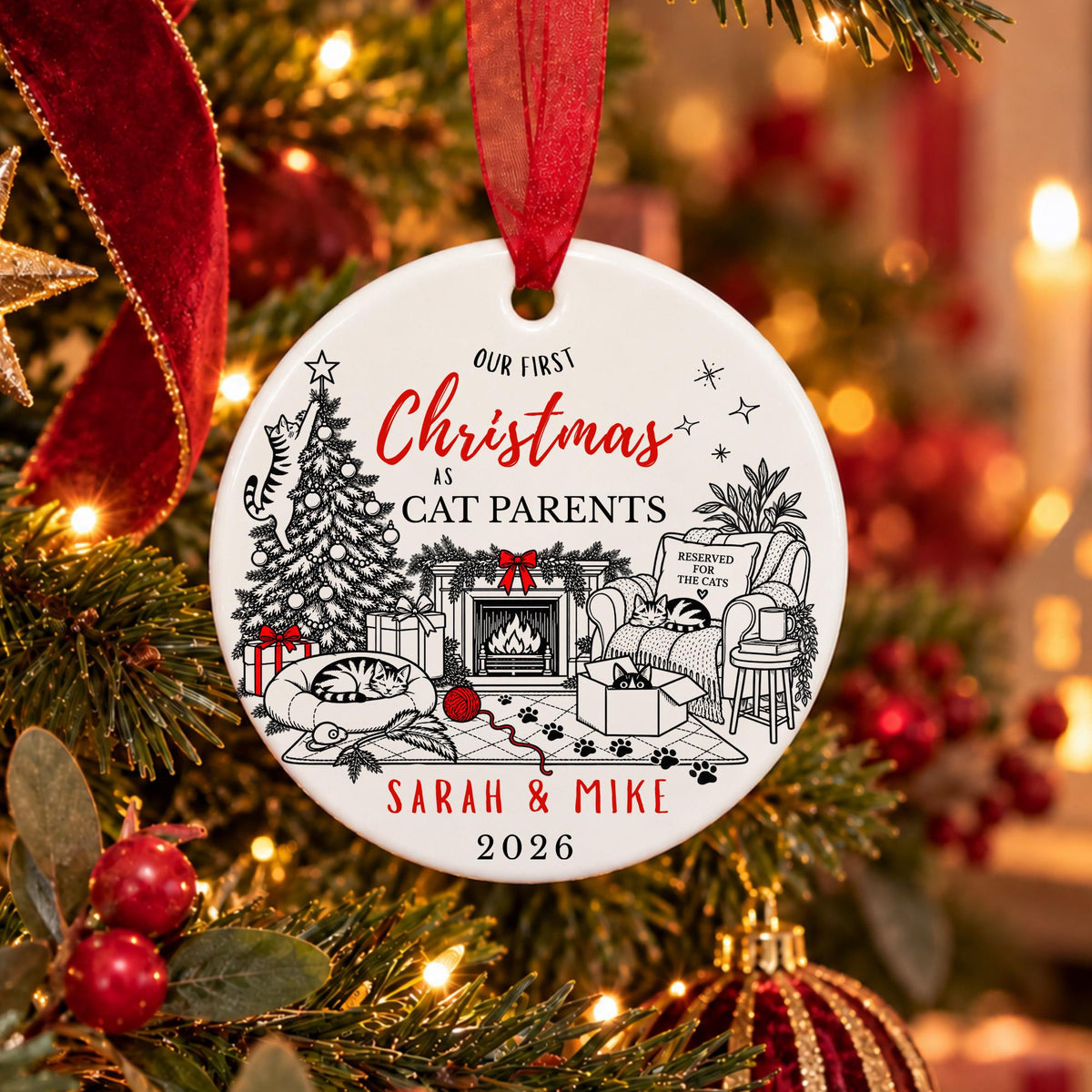 A personalized round Christmas ornament hanging on a decorated Christmas tree. The white ornament features black line art of a cozy living room with a cat on a sofa, a fireplace, a Christmas tree, and a cat sleeping on a rug, with red and black text reading 'OUR FIRST Christmas AS CAT PARENTS' and 'SARAH & MIKE 2026'.
