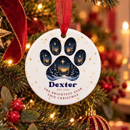 A personalized round Christmas ornament hanging on a decorated Christmas tree. The ornament is white with a large blue paw print containing a starry night sky and tree forest design, with the name 'Dexter' and the text 'THE BRIGHTEST STAR THIS CHRISTMAS' printed below.
