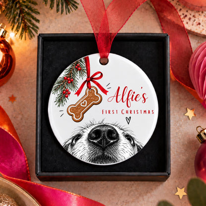 A round personalized Christmas ornament displayed in a black gift box, featuring a white background with a red ribbon, a bone-shaped ornament illustration, pine branches with red berries, and the text 'Alfie's First Christmas' above a black-and-white dog face illustration. The box is surrounded by festive holiday decorations including pine branches, lights, ribbons, and ornaments on a warm beige surface.