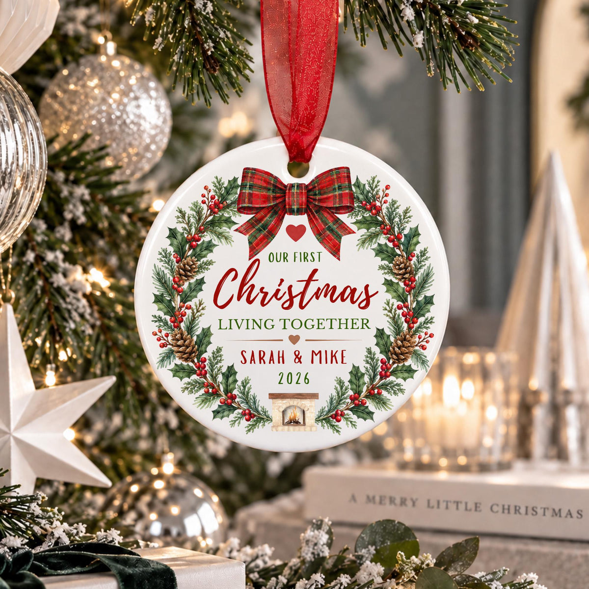 A personalized round Christmas ornament hanging from a red ribbon, featuring a festive wreath design with the text 'Our First Christmas Living Together Sarah & Mike 2026' in a decorated holiday setting with blurred Christmas lights and tree branches.
