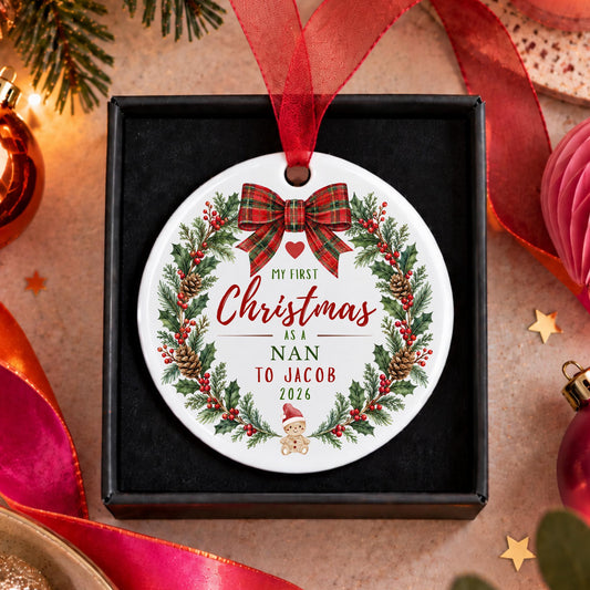 A personalized round Christmas ornament displayed in a black gift box, featuring a white background with a red ribbon, green wreath, pinecones, and the text 'My First Christmas as a Nan to Jacob 2026'.
