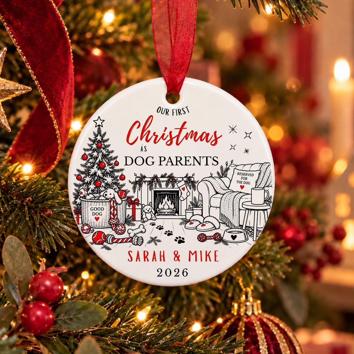 A personalized round Christmas ornament hanging on a decorated Christmas tree. The white ornament features a black line art illustration of a cozy holiday home with a dog bed, fireplace, and tree, alongside red script text reading 'Our First Christmas is Dog Parents' and the names 'Sarah & Mike' with the year '2026'.