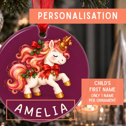 Personalized Christmas ornament featuring a white unicorn with pink and orange hair, red bows, and gold accents on a dark purple circular background. The ornament includes a red hanging ribbon and a custom name section at the bottom reading 'AMELIA', with promotional text overlays indicating personalization.